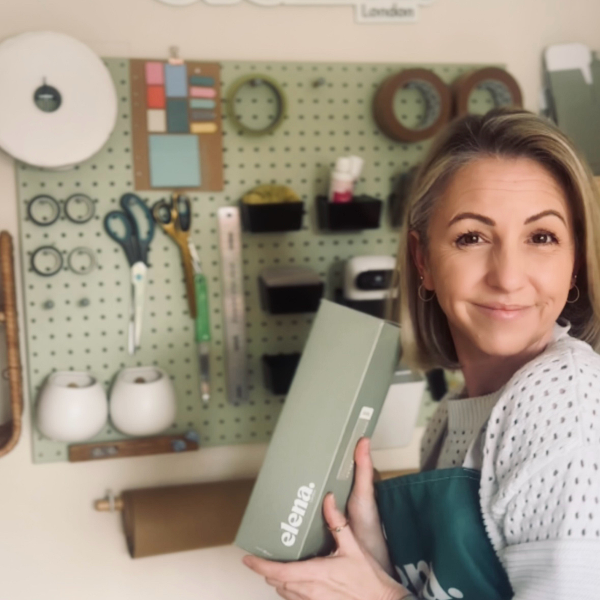 A woman holding a room spray sage green box with the brand name 'elena London.' visible on the front, standing in front of a light green pegboard filled with home fragrance supplies.
