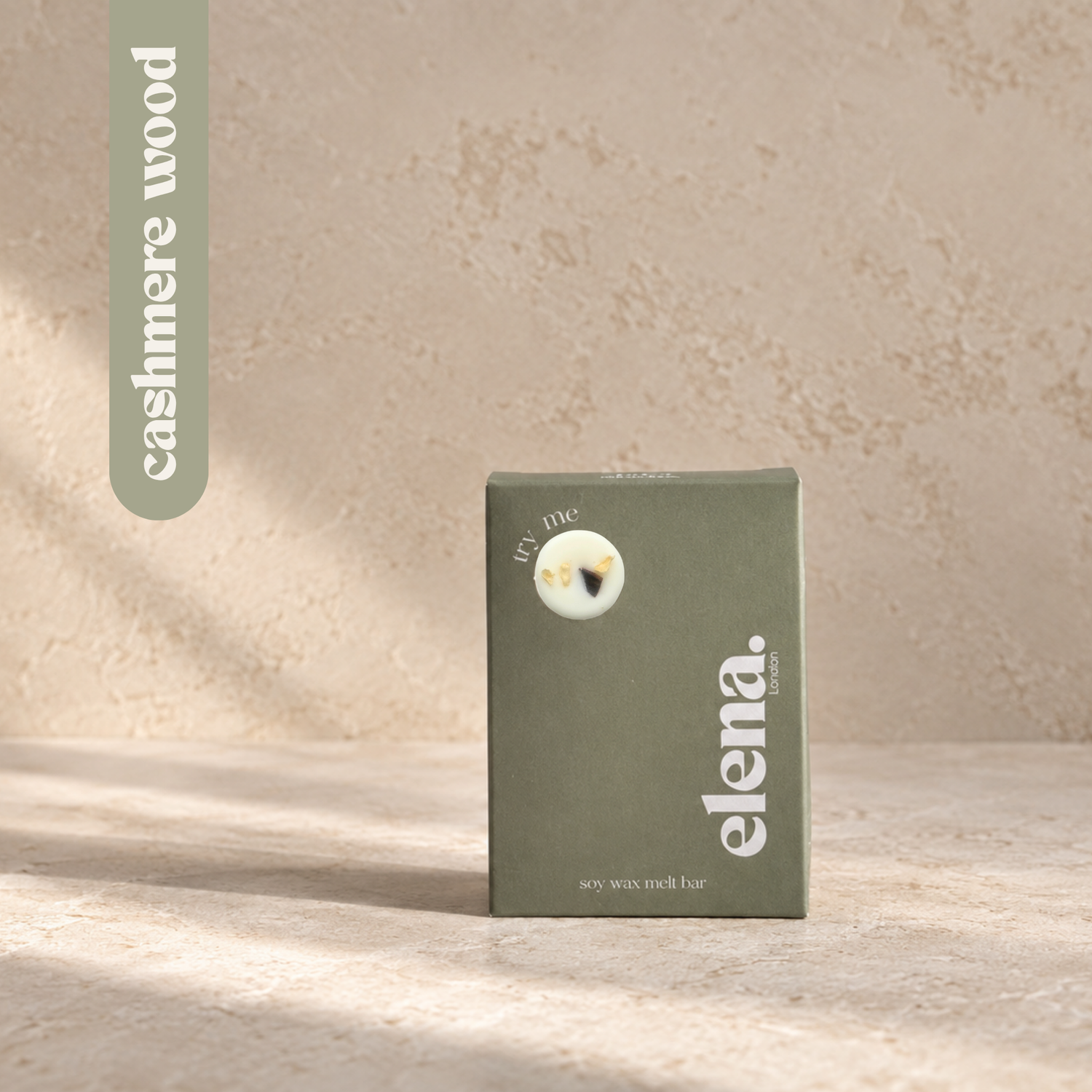 a sage green box for the elena London soy wax melt in the cashmere woof scent, standing on a textured beige surface with soft shadows.