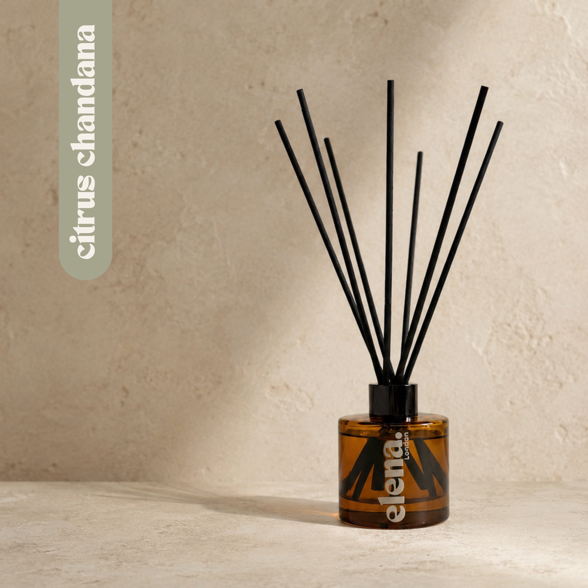 A reed diffuser bottle with black reeds, made of amber glass, on a textured beige surface against a matching beige wall. A vertical label on the left reads 'citrus chandana'.
