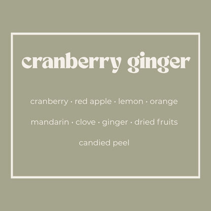 A square graphic with a muted sage green background and a thin white border. The text reads 'cranberry ginger' in large white serif letters, with smaller white text below listing scent notes: cranberry, red apple, lemon, orange, mandarin, clove, ginger, dried fruits, and candied peel.