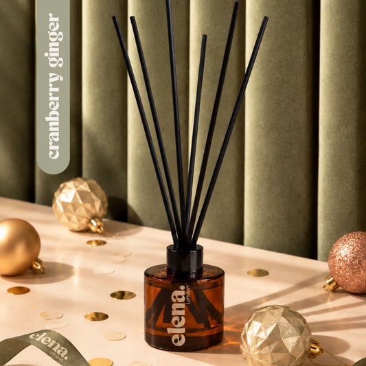 A reed diffuser bottle with black reeds, amber glass, and an elena London logo, styled on a tabletop with gold ornaments and confetti against olive green curtains. A vertical label reads 'cranberry ginger'.
