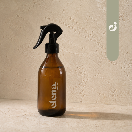 An amber glass room spray bottle with a black trigger sprayer and sage green  'elena. LONDON' branding, set against a textured beige stone background.