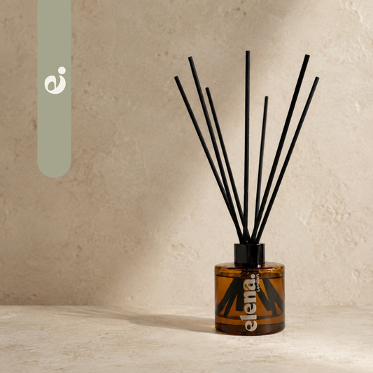 An amber glass reed diffuser bottle with black diffuser sticks, set on a textured beige surface against a matching beige wall. The brand name 'elena London' is printed vertically on the bottle. elena London signature scent, e.