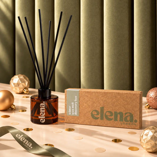 Elena London reed diffuser bottle with black reeds next to a wax melt discovery box, styled on a light surface with gold confetti, ornaments, and a ribbon against a green pleated backdrop.