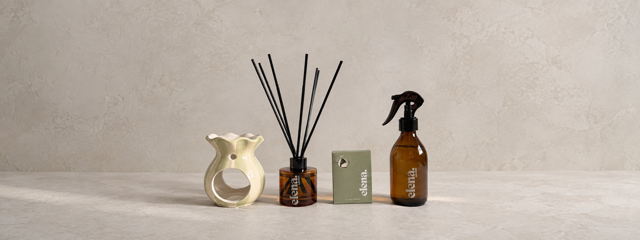 A set of four home fragrance products from the brand elena London: a sage green and cream ceramic diffuser, a reed diffuser with dark reeds, and an amber spray bottle with a black sprayer, all arranged on a light stone surface against a textured beige wall.