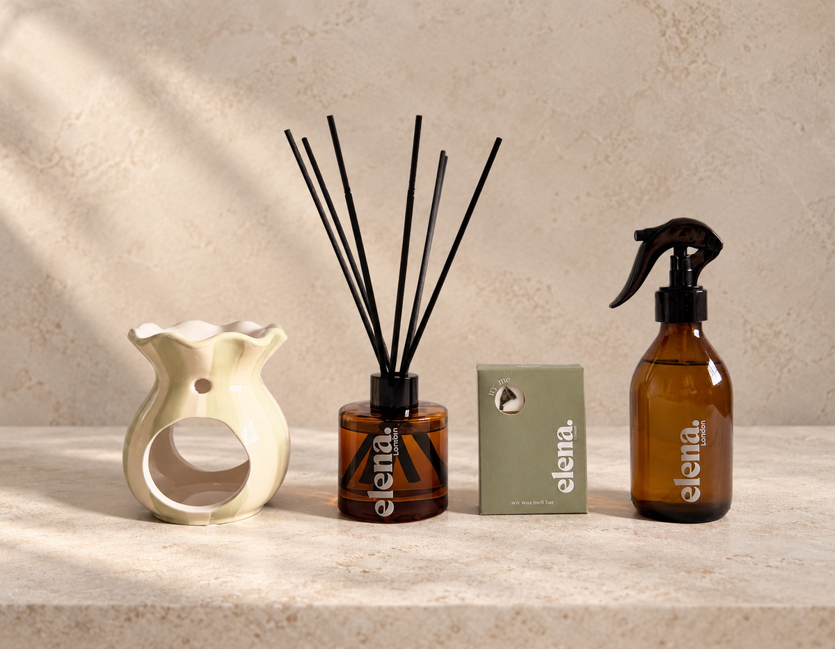A set of four elena London home fragrance products arranged on a light stone surface against a textured beige wall. The set includes a sage green and cream ceramic wax burner, a reed diffuser bottle with black reeds, a small sage green soy wax melt box, and a room spray bottle with a black trigger sprayer