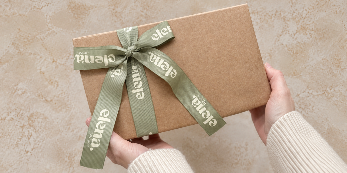 A person holds a square kraft paper gift box tied with a sage green ribbon printed with the brand name 'elena.' in white, set against a textured beige background. gifts for new homeowners. gifts for her. gifts for christmas