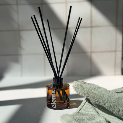 A reed diffuser with a small amber glass bottle and several black reeds, placed on a white surface with a folded sage green towel nearby and a tiled wall in the background.