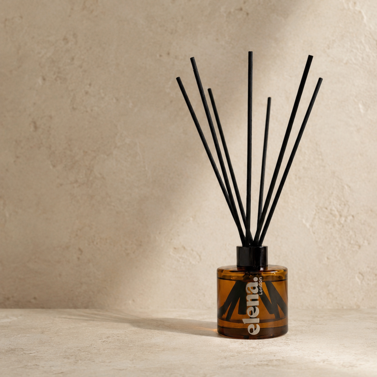 An amber glass reed diffuser bottle with black diffuser sticks, set on a textured beige surface against a matching beige wall. The brand name 'elena London' is printed vertically on the bottle.