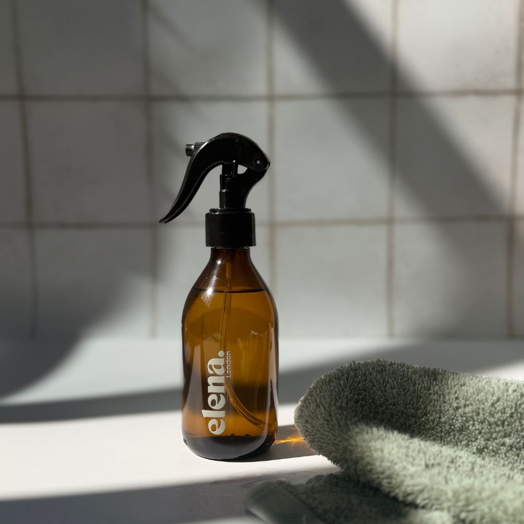 An amber glass room spray bottle with a black trigger sprayer and the brand name 'elena London' printed in sage green on the front, set on a white tiled surface with a folded green towel nearby and strong diagonal shadows.