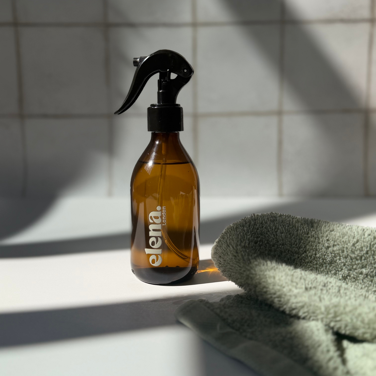 An amber glass spray bottle with a black trigger sprayer and white 'elena London' branding, set on a white surface next to a folded sage green towel with strong diagonal sunlight and tiled background.