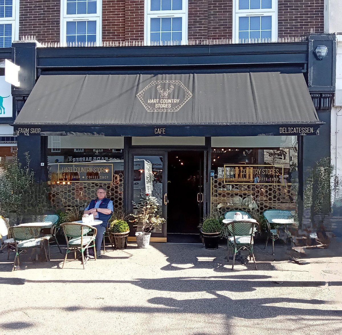 A storefront scene for Hart Country Coffee Shop and gift shop in Haywards heath. with a dark awning, brick building facade, outdoor seating, and a seated person visible through the window.