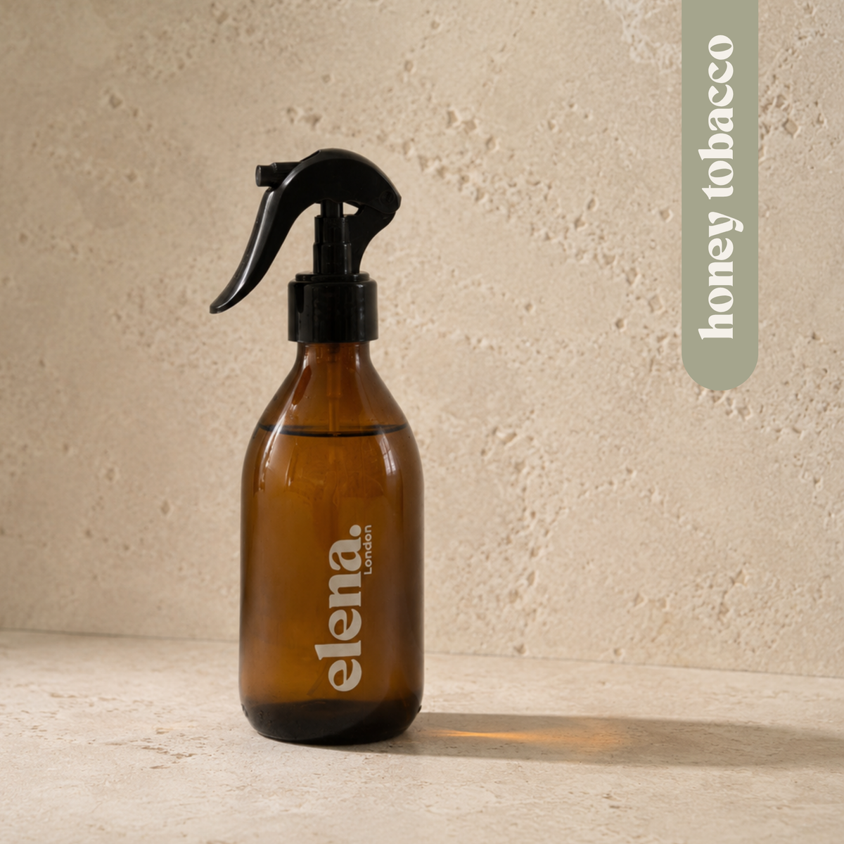 An amber glass room spray bottle with a black trigger sprayer and sage green 'elena. LONDON' branding, set against a textured beige stone background. honey tobacco scent