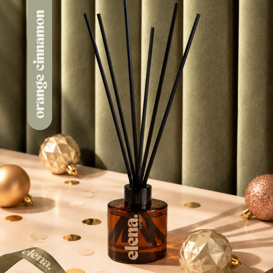 An amber glass reed diffuser bottle with black reeds and the elena London logo, styled on a tabletop with gold ornaments and confetti against a green curtain background. A vertical label reads 'orange cinnamon'.