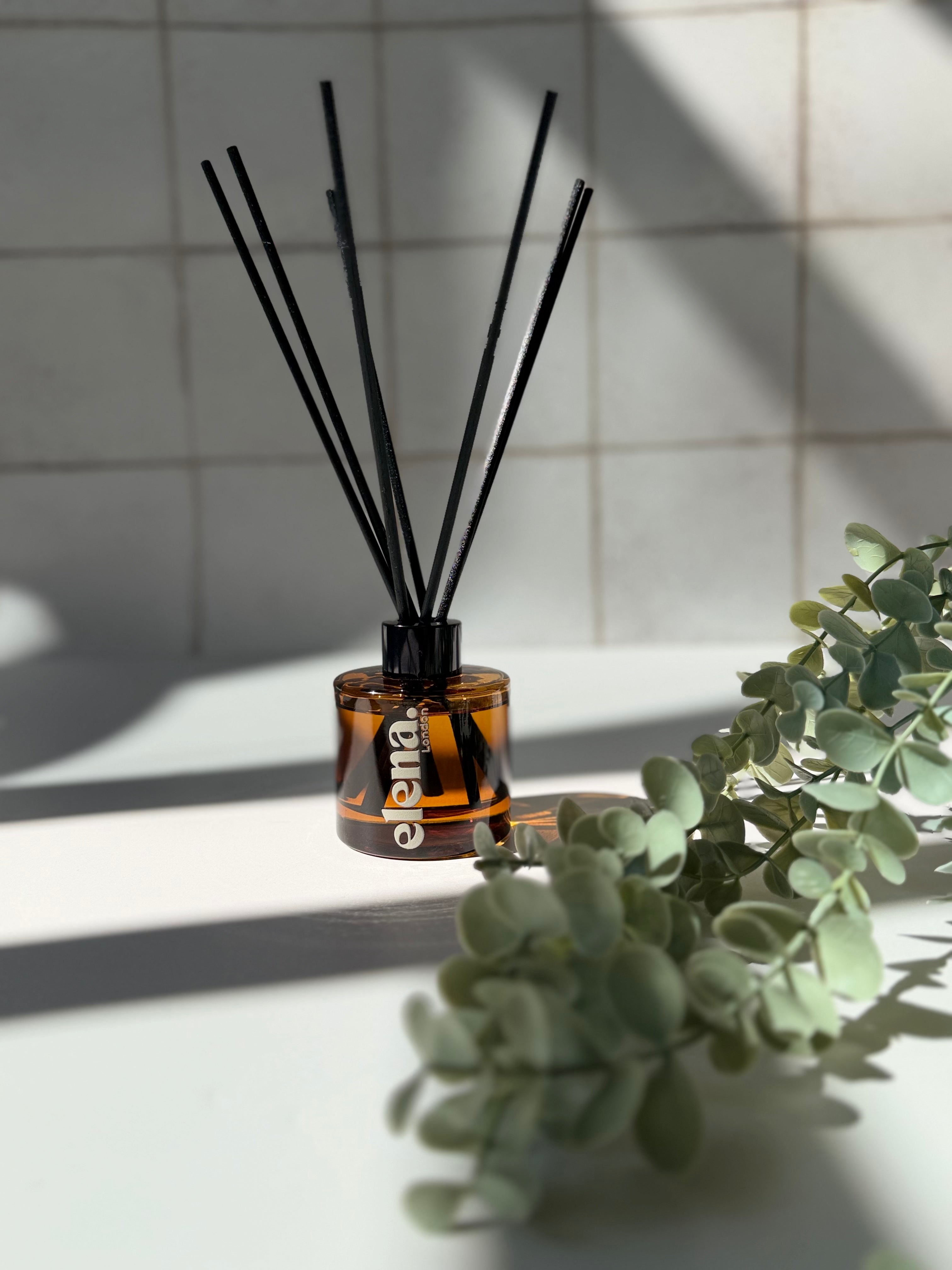 An amber glass reed diffuser bottle with black diffuser sticks, placed on a white surface with soft shadows and green eucalyptus leaves in the foreground.