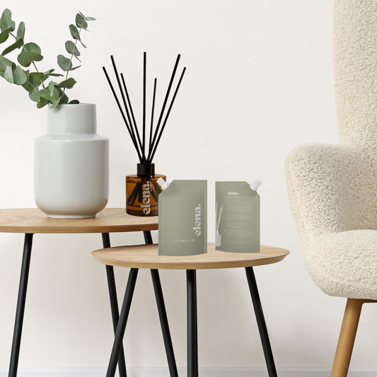 Two sage green reed diffuser refill pouches sit on a small wooden side table in a styled interior setting with a cream boucle armchair, a white ceramic vase with greenery, and a reed diffuser from elena London