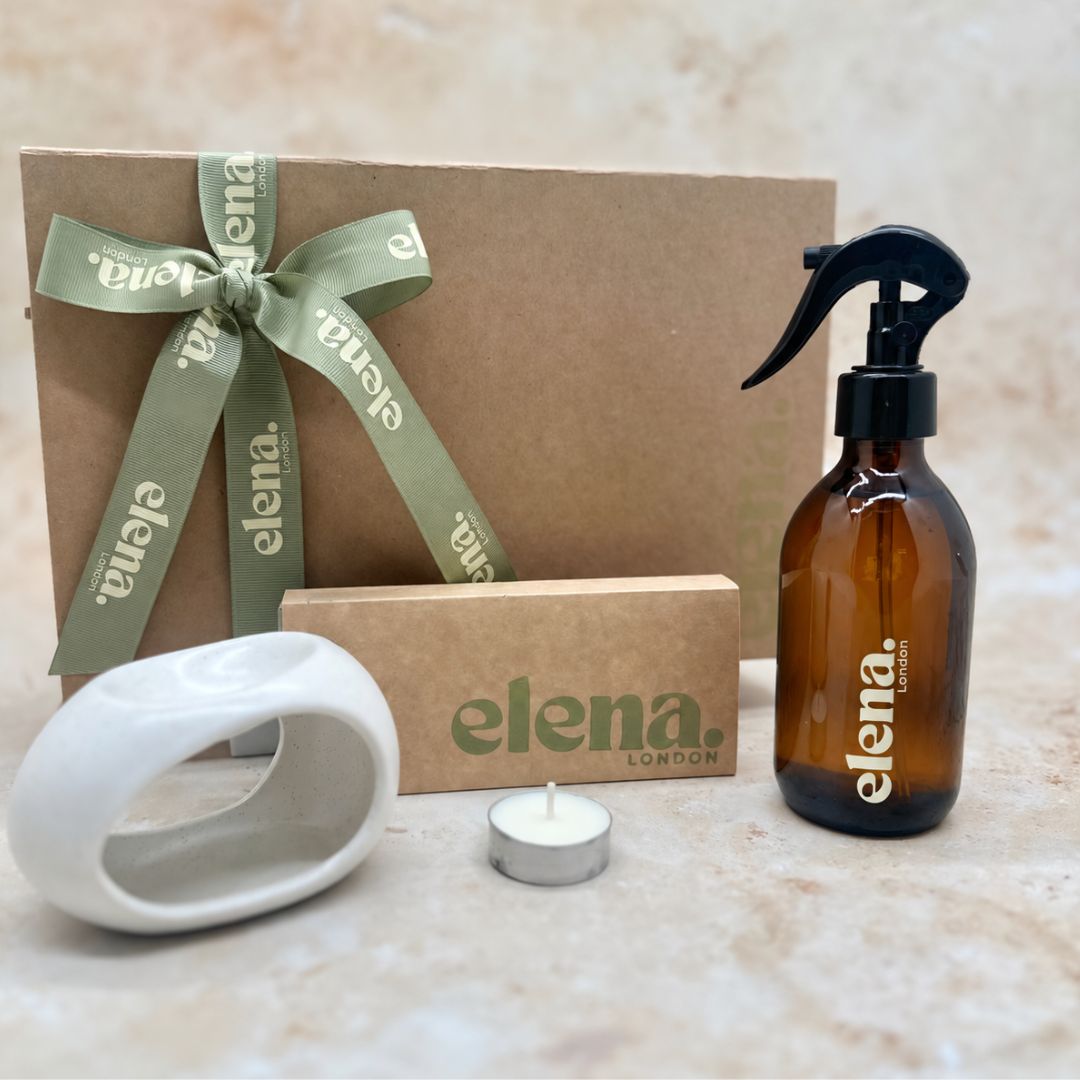 A gift set from Elena London featuring an amber glass room spray bottle with a black sprayer, a brown kraft gift box tied with an sage green ribbon, a wax melt discovery box, a white ceramic wax burner, and a tea light candle on a beige stone surface.