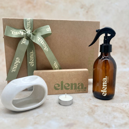 A gift set from Elena London featuring an amber glass room spray bottle with a black sprayer, a brown kraft gift box tied with an sage green ribbon, a wax melt discovery box, a white ceramic wax burner, and a tea light candle on a beige stone surface.