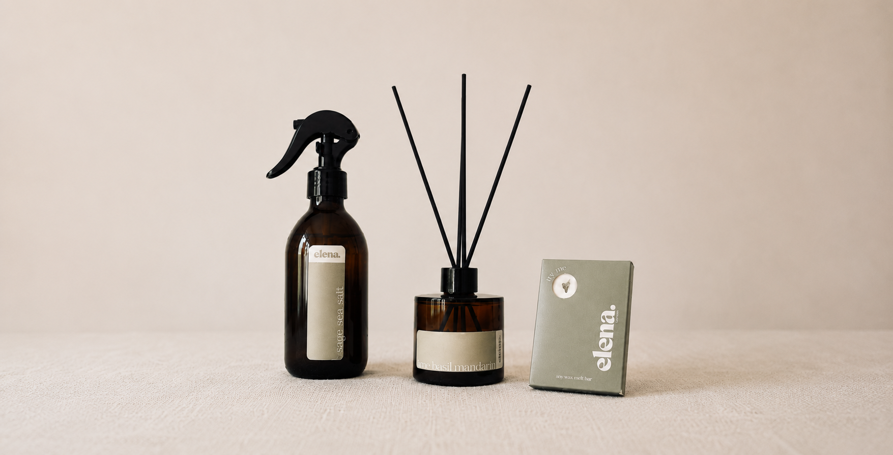 Three minimalist home fragrance items arranged on a light beige surface against a warm beige background: an sage sea salt spray bottle with a black sprayer, a lime basil mandarinreed diffuser bottle with black reeds, and a small sage green box with a visible 'elena' logo with a wax melt in.