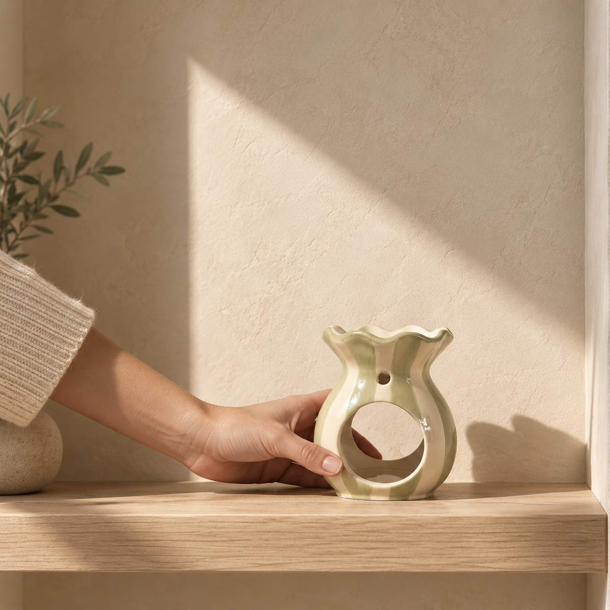 A hand in a beige knit sweater holds a ceramic wax melt burner with a wavy rim and central cutout, placed on a light wood shelf against a textured beige wall with warm sunlight and shadow.