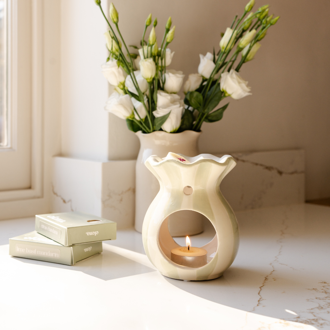 Decorative sage green wax burner with a tealight and a vase of flowers on a marble surface.