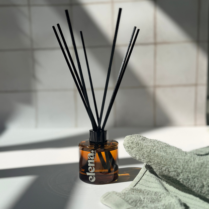A reed diffuser bottle with black reeds, placed on a white surface with a folded sage green towel beside it and a tiled wall in the background. The brand name 'elena London' is visible on the bottle.