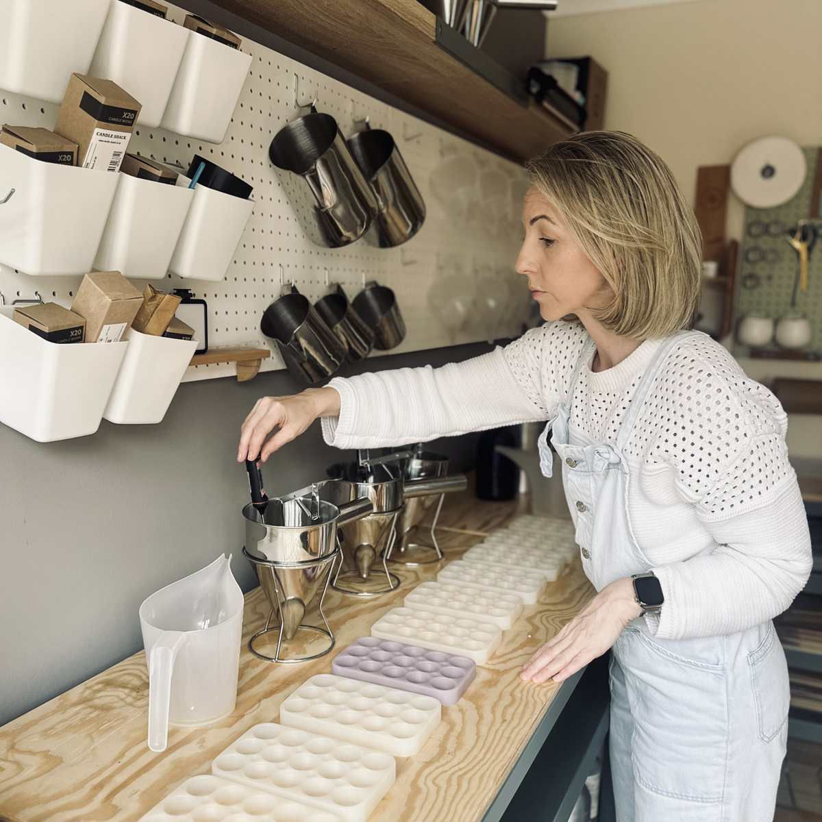A woman in a white long-sleeve top and light denim overalls stands at a wooden counter in a home fragrance studio, stirring metal containers filled with melted wax while several white oval-shaped wax melt moulds are laid out on the counter.