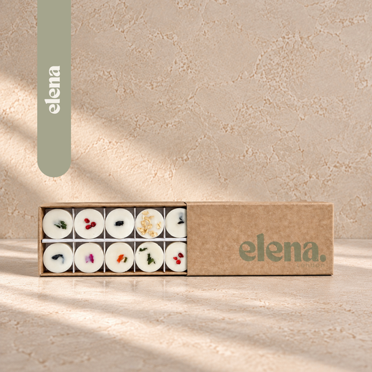 An open kraft paper box containing ten wax melts, each decorated with dried botanicals, shown on a textured beige surface with soft diagonal shadows. The brand name 'elena London