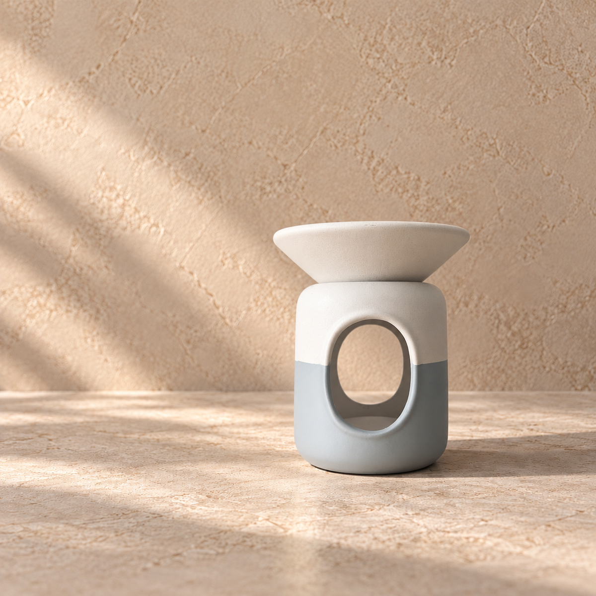 A ceramic oil burner warmer with a white top bowl and a two-tone base in white and light grey, set on a textured beige stone surface with soft diagonal shadows.