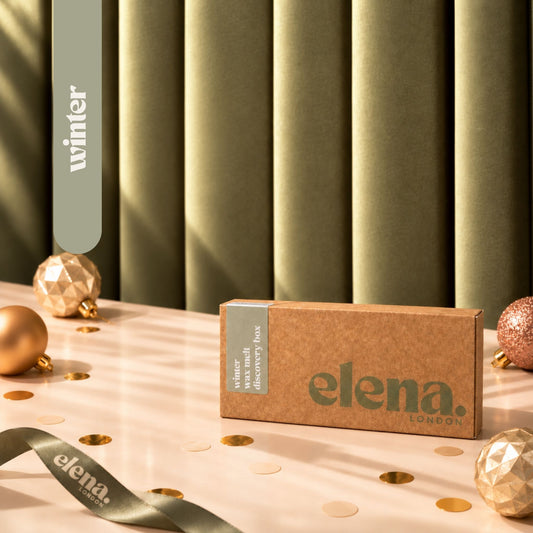 A kraft wax melt discovery box labeled 'elena London winter winter discovery box' sits on a light pink surface with gold confetti, decorative christmas ornaments, and a matching ribbon, against a green pleated backdrop.